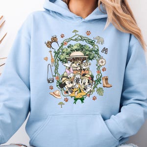 Peut inclure: Sweat &agrave; capuche bleu clair avec un motif sur le th&egrave;me du safari. Le design comprend des personnages de dessins anim&eacute;s, un v&eacute;hicule de safari et des ic&ocirc;nes de safari telles qu'une girafe, un &eacute;l&eacute;phant et des jumelles. Le motif est entour&eacute; d'une couronne de feuilles et d'empreintes de pattes.