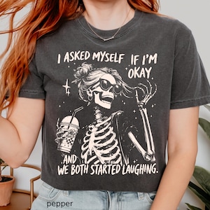 May include: Dark gray t-shirt featuring a skeleton wearing sunglasses and holding a drink. The shirt has the text "I asked myself if I'm okay and we both started laughing."