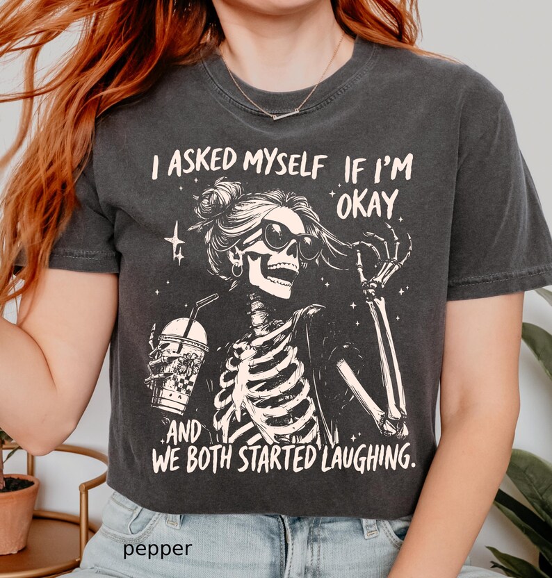 May include: Dark gray t-shirt featuring a skeleton wearing sunglasses and holding a drink. The shirt has the text "I asked myself if I'm okay and we both started laughing."