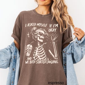 May include: Espresso-colored t-shirt featuring a skeleton wearing sunglasses and holding a drink. The graphic includes the text "I asked myself if I'm okay and we both started laughing."