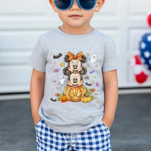 Puede incluir: Una camiseta gris de manga corta con un dise&ntilde;o de Halloween. El dise&ntilde;o incluye personajes de dibujos animados apilados, una calabaza, fantasmas, murci&eacute;lagos y estrellas. La camiseta la lleva un ni&ntilde;o con gafas de sol azules y pantalones cortos a cuadros azules y blancos.