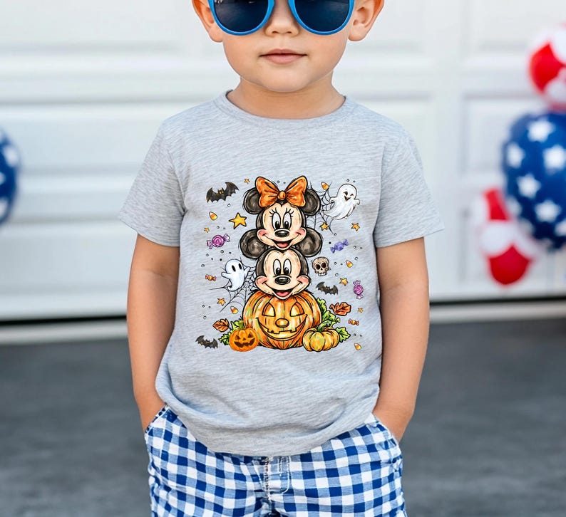 Puede incluir: Una camiseta gris de manga corta con un dise&ntilde;o de Halloween. El dise&ntilde;o incluye personajes de dibujos animados apilados, una calabaza, fantasmas, murci&eacute;lagos y estrellas. La camiseta la lleva un ni&ntilde;o con gafas de sol azules y pantalones cortos a cuadros azules y blancos.