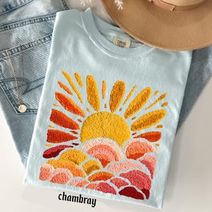 May include: Light blue t-shirt with an embroidered sunset design in shades of yellow, orange, and red. The word "chambray" is printed at the bottom. The shirt is folded on top of blue jeans, with a necklace and a brown hat in the background.