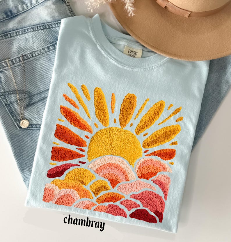May include: Light blue t-shirt with an embroidered sunset design in shades of yellow, orange, and red. The word "chambray" is printed at the bottom. The shirt is folded on top of blue jeans, with a necklace and a brown hat in the background.