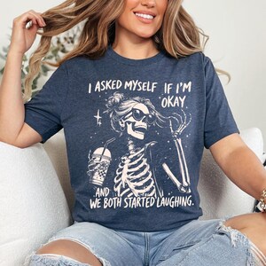 May include: Navy blue t-shirt with a skeleton graphic wearing sunglasses and holding a drink. The text reads, "I asked myself if I'm okay. And we both started laughing."