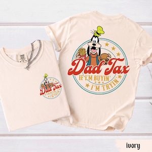 May include: Ivory-colored t-shirts featuring a cartoon character holding a burger and fries, with the text "Dad Tax" in red script. The design is repeated on the front pocket and back of the shirts. The shirts have a round neckline and short sleeves.