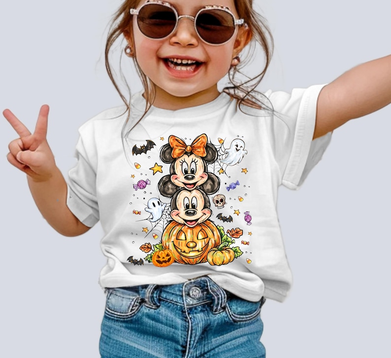 Puede incluir: Camiseta blanca con un dise&ntilde;o de Halloween de Mickey y Minnie Mouse apilados sobre una calabaza tallada. El dise&ntilde;o incluye fantasmas, murci&eacute;lagos, estrellas y caramelos. La camiseta la lleva un ni&ntilde;o.