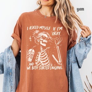 May include: Rust-colored t-shirt featuring a skeleton graphic with sunglasses and a drink, along with the text "I asked myself if I'm okay, and we both started laughing."