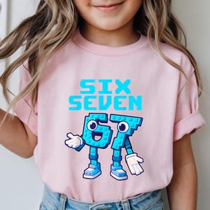May include: A light pink t-shirt featuring the text "SIX SEVEN" in pixelated blue letters. Below the text are the numbers "67" designed as cartoon characters with blue bodies, white gloves, and blue shoes. The shirt is for children.