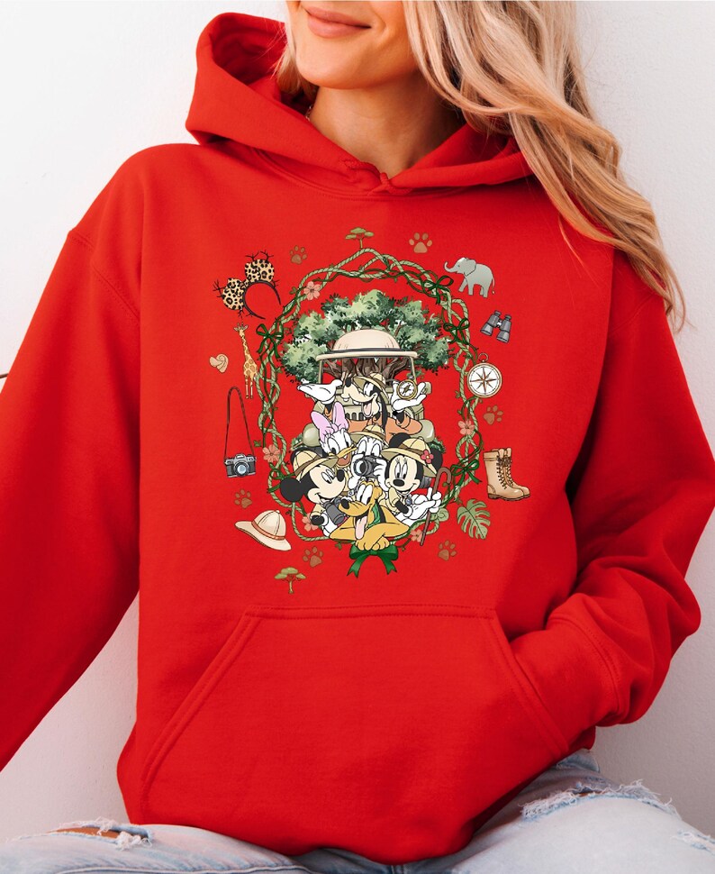 Peut inclure: Sweat &agrave; capuche rouge avec un motif de safari. Le motif comprend un chapeau de safari, des jumelles, une boussole et un appareil photo. Le motif est entour&eacute; d'une couronne de feuilles et d'animaux de safari.