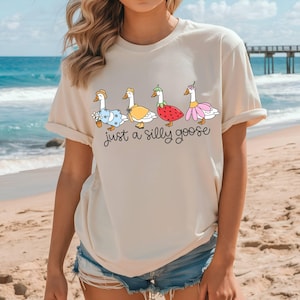 May include: Beige t-shirt featuring a whimsical illustration of four geese dressed in various outfits, including a straw hat, a yellow dress, a strawberry costume, and a pink flower dress. The text "just a silly goose" is written below the geese.