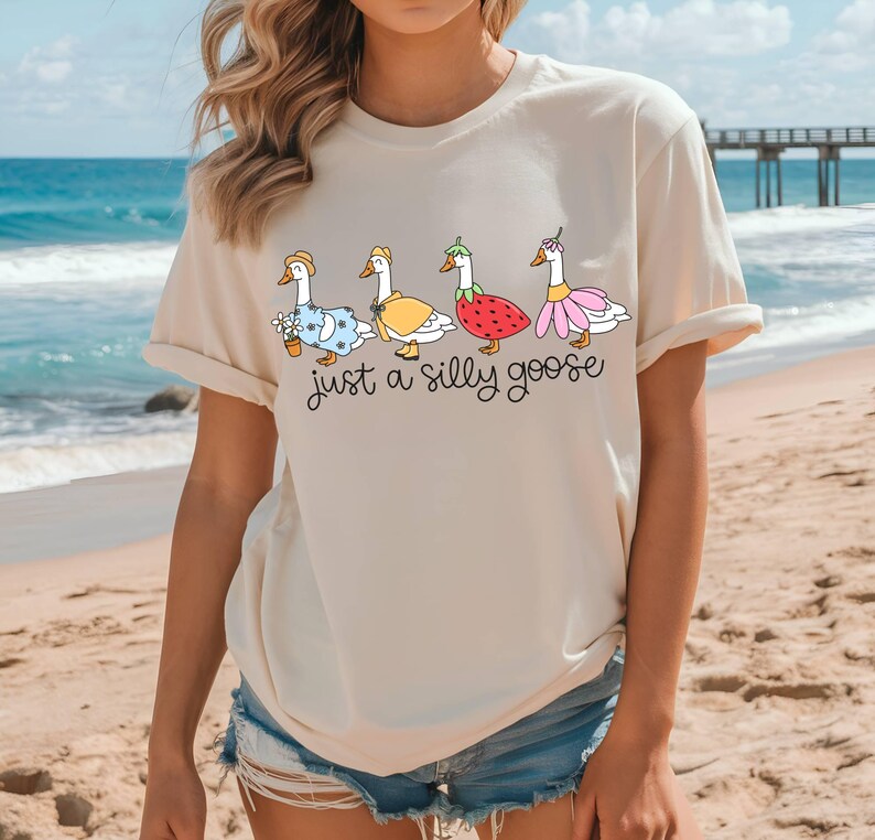 May include: Beige t-shirt featuring a whimsical illustration of four geese dressed in various outfits, including a straw hat, a yellow dress, a strawberry costume, and a pink flower dress. The text "just a silly goose" is written below the geese.