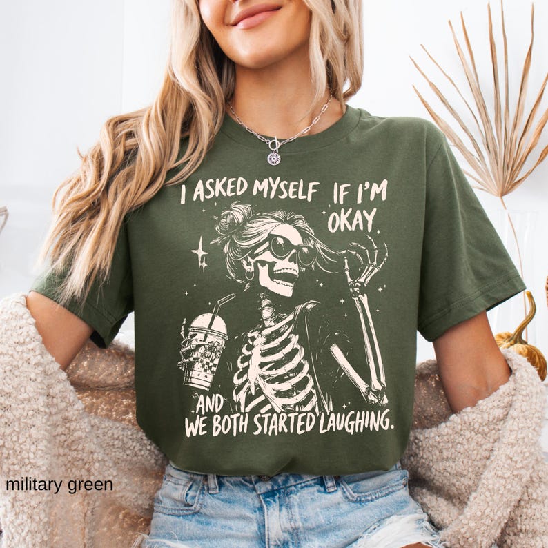 May include: Military green t-shirt featuring a skeleton graphic wearing sunglasses and holding a drink. The text on the shirt reads, "I asked myself if I'm okay, and we both started laughing."