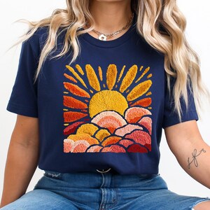 May include: Navy blue t-shirt featuring an embroidered design of a yellow sun with orange and red rays and clouds. The design is centered on the shirt. The shirt is a short-sleeved crew neck.