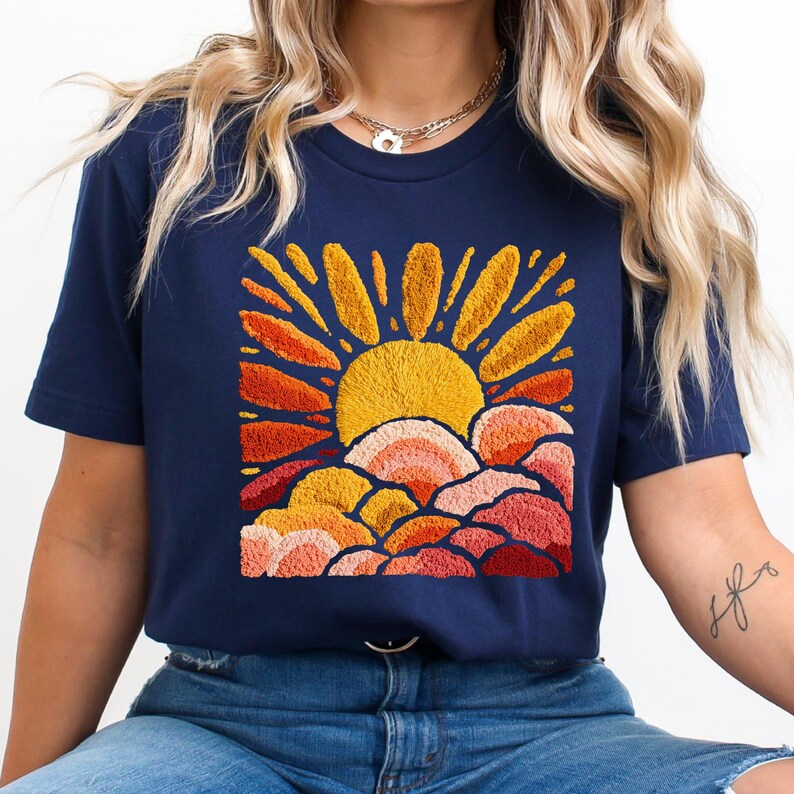 May include: Navy blue t-shirt featuring an embroidered design of a yellow sun with orange and red rays and clouds. The design is centered on the shirt. The shirt is a short-sleeved crew neck.