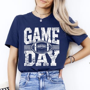Puede incluir: Camiseta azul marino con las palabras "GAME DAY" en letras blancas y un gr&aacute;fico de f&uacute;tbol americano. La camiseta es de cuello redondo y tiene mangas cortas. La camiseta es de material suave.
