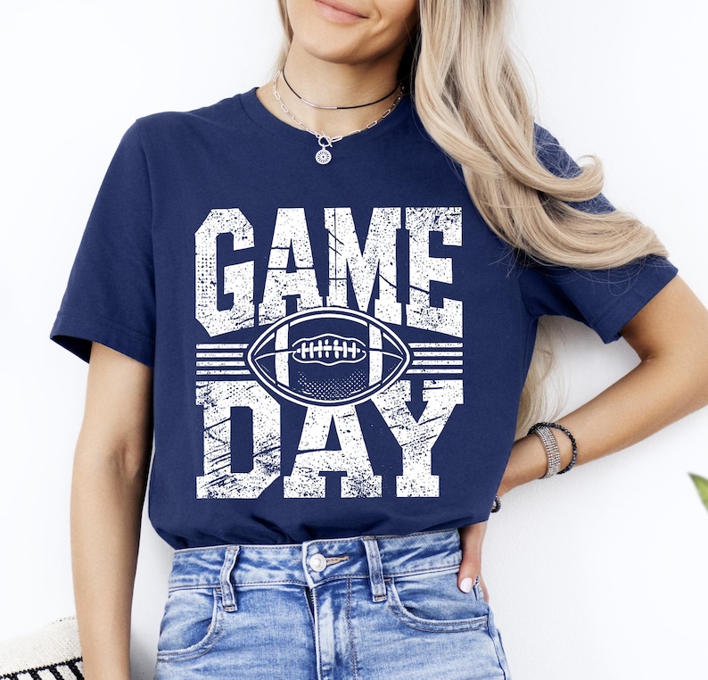 Puede incluir: Camiseta azul marino con las palabras "GAME DAY" en letras blancas y un gr&aacute;fico de f&uacute;tbol americano. La camiseta es de cuello redondo y tiene mangas cortas. La camiseta es de material suave.