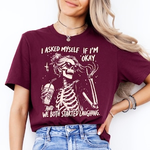 May include: Burgundy t-shirt with a skeleton graphic wearing sunglasses and holding a drink. The text reads, "I asked myself if I'm okay, and we both started laughing."