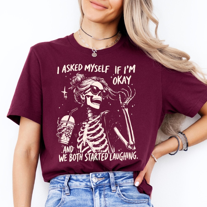 May include: Burgundy t-shirt with a skeleton graphic wearing sunglasses and holding a drink. The text reads, "I asked myself if I'm okay, and we both started laughing."