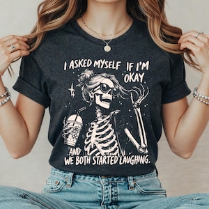 May include: Dark gray t-shirt featuring a skeleton graphic wearing sunglasses and holding a drink. The text on the shirt reads, "I asked myself if I'm okay. And we both started laughing."