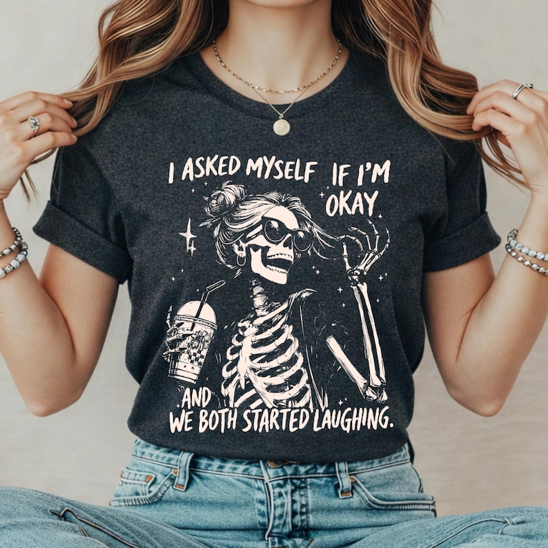 May include: Dark gray t-shirt featuring a skeleton graphic wearing sunglasses and holding a drink. The text on the shirt reads, "I asked myself if I'm okay. And we both started laughing."