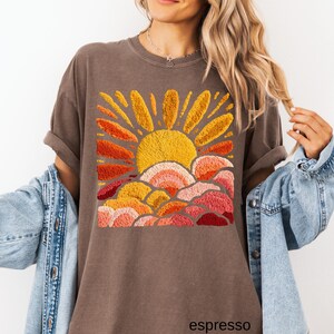 May include: Espresso-colored t-shirt featuring an embroidered sun design in shades of yellow, orange, and red. The shirt has a relaxed fit and rolled sleeves. The word "espresso" is printed at the bottom.