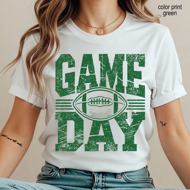 May include: A white short-sleeved t-shirt featuring a distressed green graphic print. The design displays the text "GAME DAY" in a bold, block font with a football illustration centered between the words. The text "color print green" is visible.