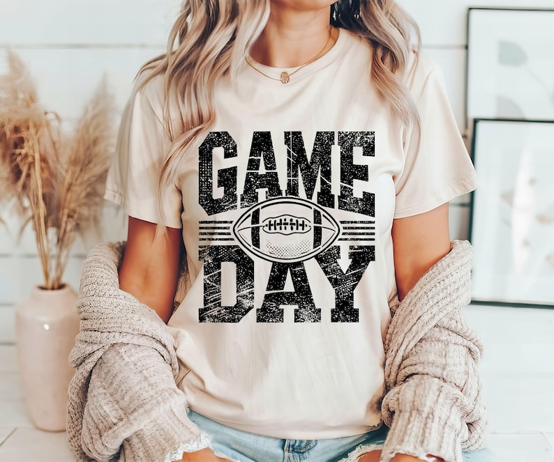 Puede incluir: Camiseta color crema con las palabras "GAME DAY" en letras negras desgastadas, con un gr&aacute;fico de f&uacute;tbol americano. La camiseta es de estilo casual, adecuada para el d&iacute;a del partido o el uso diario.