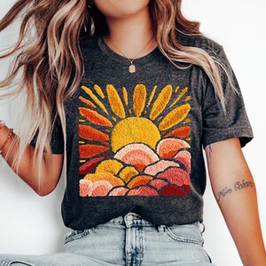 May include: Dark gray t-shirt featuring an embroidered design of a sun rising over layered clouds. The sun is yellow and orange, with rays extending outwards. The clouds are in shades of orange, pink, and red.