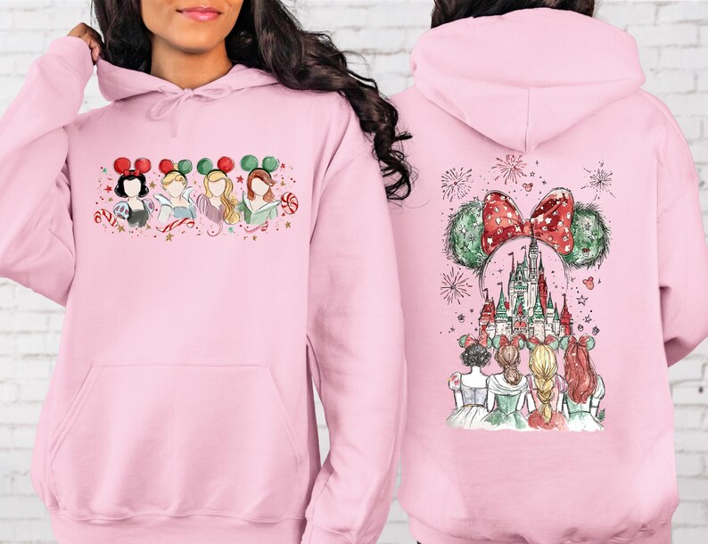 May include: A light pink hoodie featuring a watercolor design of Disney princesses. The front shows four princesses with Mickey Mouse ears. The back displays a castle, fireworks, and more princesses with Mickey Mouse ears.