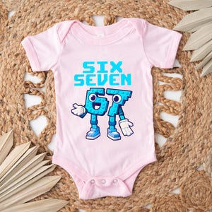 May include: A light pink baby onesie with the words "SIX SEVEN" in pixelated blue letters. Below the text are cartoon figures of the numbers 6 and 7, also in blue, with white gloves and shoes. The onesie has a snap closure.