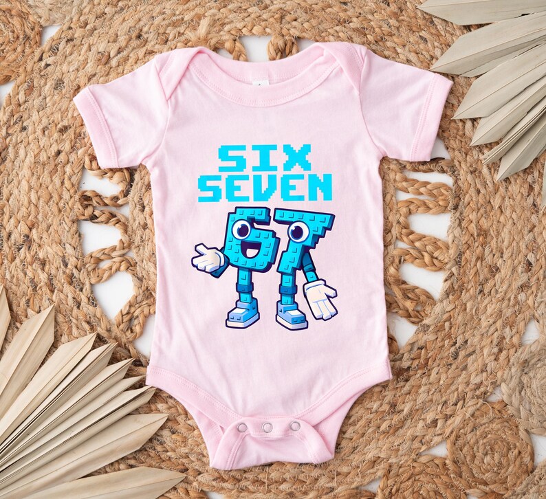 May include: A light pink baby onesie with the words "SIX SEVEN" in pixelated blue letters. Below the text are cartoon figures of the numbers 6 and 7, also in blue, with white gloves and shoes. The onesie has a snap closure.