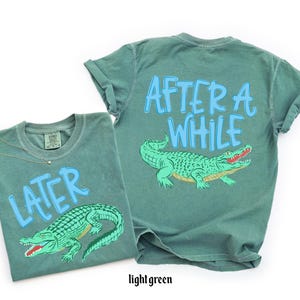 May include: Two light green t-shirts with alligator graphics. One shirt says "LATER" and the other says "AFTER A WHILE" in light blue. The alligators are green with red mouths and yellow bellies.