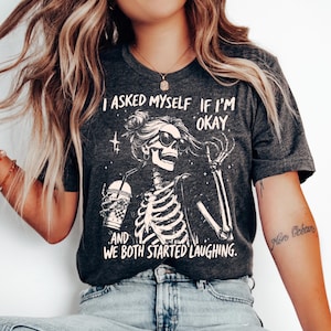 May include: Dark gray t-shirt featuring a skeleton wearing sunglasses and holding a drink. The shirt has the text "I asked myself if I'm okay and we both started laughing." The graphic is in white.