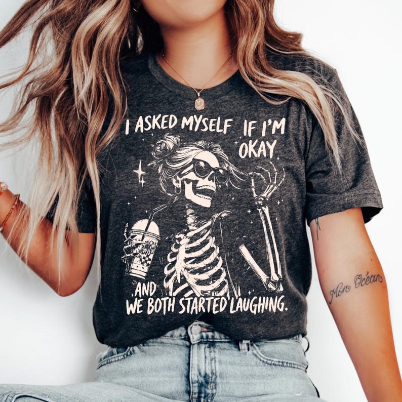 May include: Dark gray t-shirt featuring a skeleton wearing sunglasses and holding a drink. The shirt has the text "I asked myself if I'm okay and we both started laughing." The graphic is in white.
