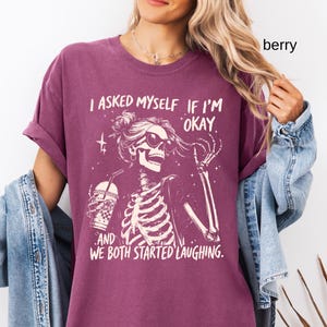 May include: Berry-colored t-shirt featuring a skeleton graphic wearing sunglasses and holding a drink. The text reads, "I asked myself if I'm okay, and we both started laughing." The shirt has rolled-up sleeves and a relaxed fit.