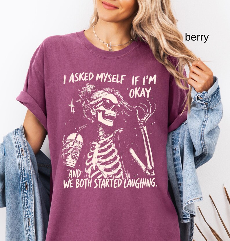May include: Berry-colored t-shirt featuring a skeleton graphic wearing sunglasses and holding a drink. The text reads, "I asked myself if I'm okay, and we both started laughing." The shirt has rolled-up sleeves and a relaxed fit.