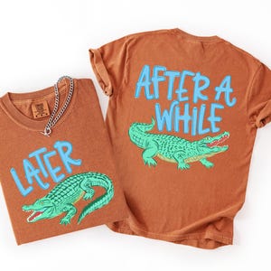 May include: Two rust-colored t-shirts with the phrases "LATER" and "AFTER A WHILE" in light blue, each featuring a green alligator illustration. One shirt has a silver chain necklace. The shirts are likely part of a matching set.