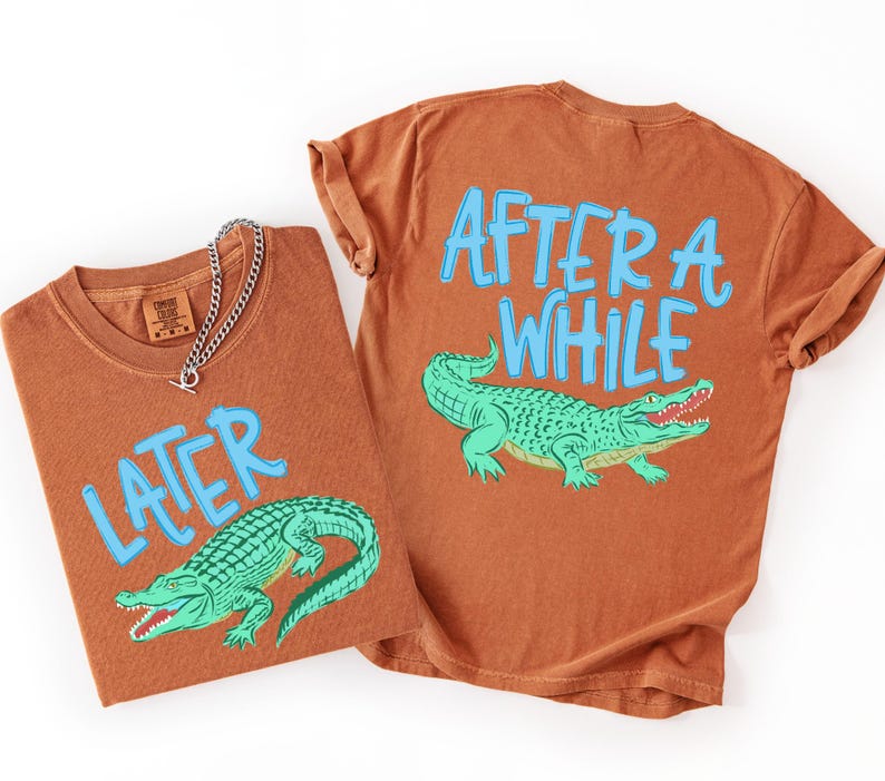 May include: Two rust-colored t-shirts with the phrases "LATER" and "AFTER A WHILE" in light blue, each featuring a green alligator illustration. One shirt has a silver chain necklace. The shirts are likely part of a matching set.