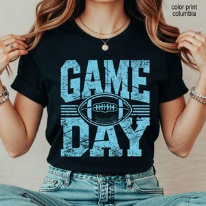 May include: A black short-sleeve t-shirt featuring the text "GAME DAY" in a distressed light blue font. Centered between the words is a graphic of an American football. The text "color print columbia" is visible in the upper right corner.