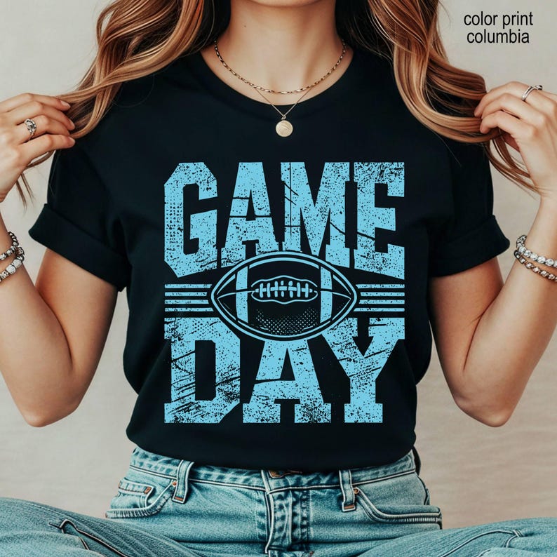 May include: A black short-sleeve t-shirt featuring the text "GAME DAY" in a distressed light blue font. Centered between the words is a graphic of an American football. The text "color print columbia" is visible in the upper right corner.