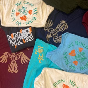 May include: A collection of various t-shirts in different colors, including white, navy blue, and teal. Some shirts feature the text "WE BUILD WE BURN WE GROW" around a floral design. Other shirts have graphic designs or text like "UN10" and a stylized bird.