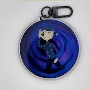 Puede incluir: Un llavero redondo con borde plateado, que presenta una ilustración de dibujos animados de una niña con un suéter azul con estrellas blancas, sobre un fondo arremolinado azul y morado. Tiene un cierre negro.