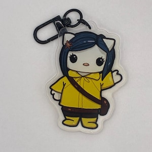 Puede incluir: Un llavero acrílico transparente con un personaje de dibujos animados con orejas de gato blancas, cabello azul oscuro y un impermeable amarillo. El personaje tiene una bandolera marrón y está saludando. El llavero tiene un cierre negro.