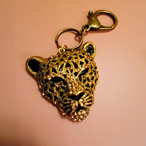 May include: A gold-tone keychain featuring a detailed, black and gold jeweled leopard head charm. The charm is attached to a gold-tone split ring and a lobster claw clasp.