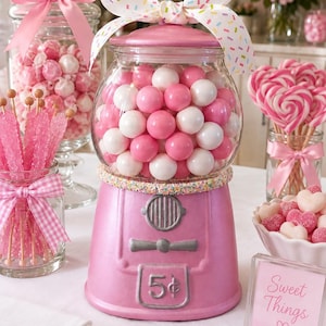 May include: A pink gumball machine with a glass globe filled with pink and white gumballs, topped with a white bow featuring colorful sprinkles. The base is pink with a silver coin slot marked 5 cents, a silver dial, and a silver dispensing lever.