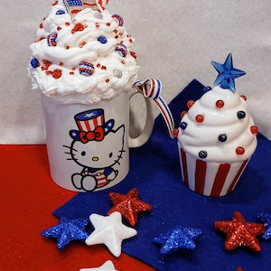 May include: A white ceramic mug featuring a Hello Kitty design wearing a red, white and blue hat. The mug is filled with whipped cream and red, white and blue sprinkles. A small white cupcake with red, white and blue frosting and sprinkles sits next to the mug. Both are on a blue surface with white, red and blue glitter stars.