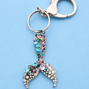 Op de afbeelding: Een zilverkleurige metalen sleutelhanger met een bedel in de vorm van een zeemeerminstaart, ingelegd met veelkleurige strass-steentjes in tinten blauw, paars, roze en transparant. De bedel zit aan een ring en een karabijnsluiting bevestigd.