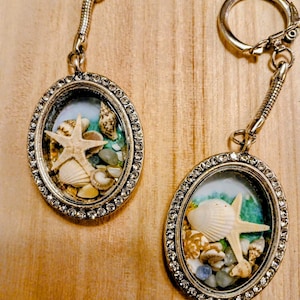 May include: Two oval-shaped keychains featuring silver-toned metal frames encrusted with small clear rhinestones. Each keychain contains a miniature display of various seashells, a small starfish, and tiny blue stones set against a white background.