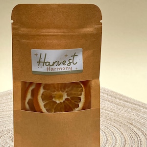 Peut inclure: Un sac en papier brun avec une fen&ecirc;tre transparente montrant des tranches d'orange s&eacute;ch&eacute;es. Le sac porte une &eacute;tiquette blanche avec le texte "Harvest Harmony" en noir.